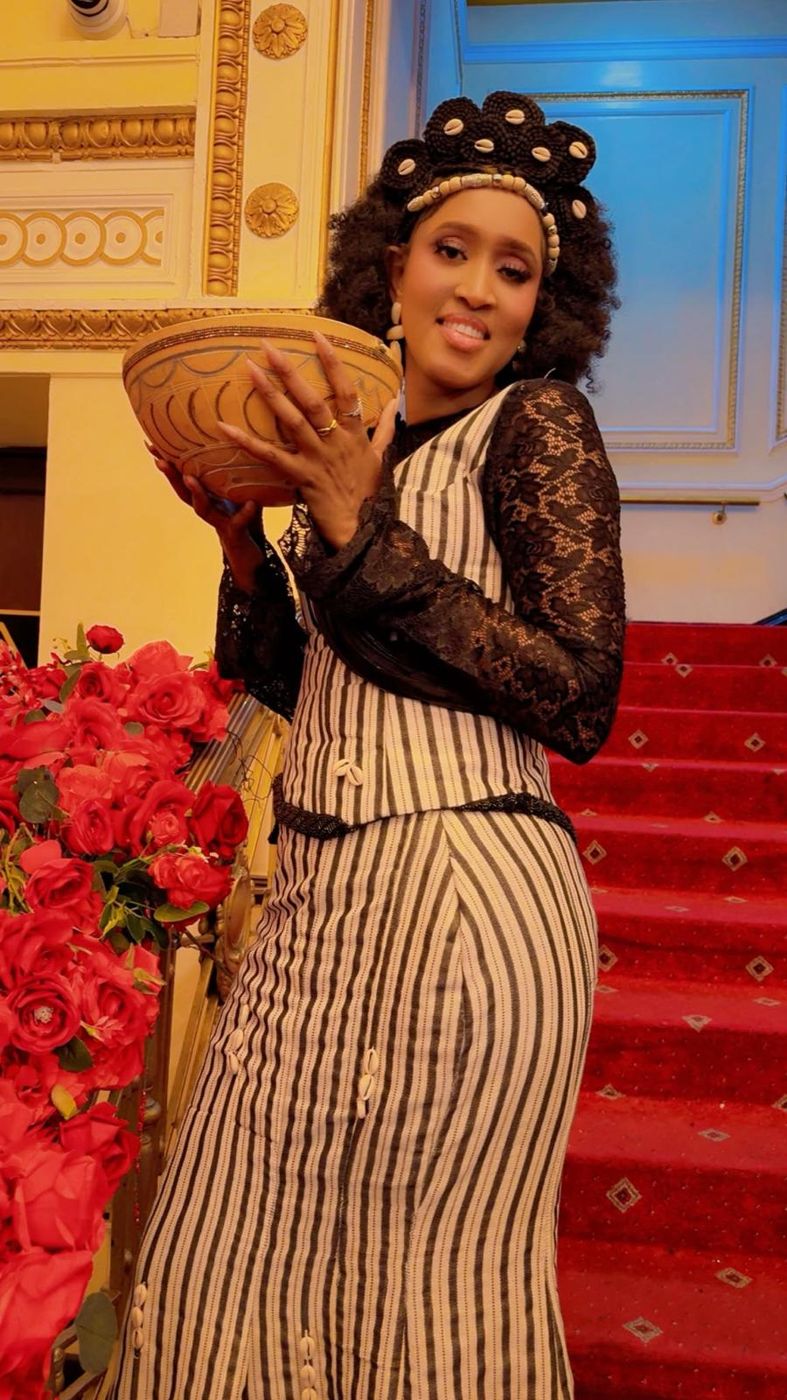A member in traditional dress and headpiece carrying a woven calabash bowl